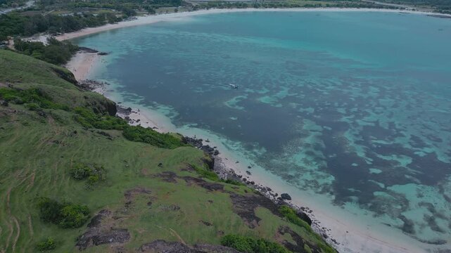 Aerial view bukit merese ridge with turquoise ocean and pink sand beach, sweeping green slopes, rocky cliffs, crystal waves breaking on shore, remote island coastline, dramatic horizon