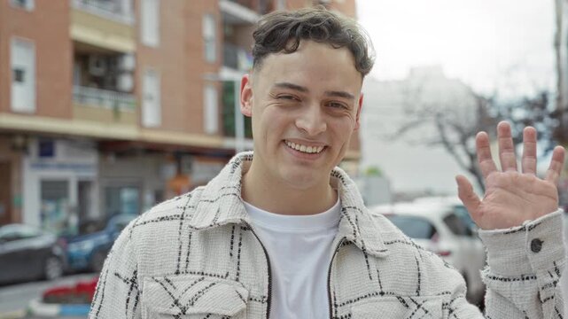 Man smiling and waving an open right hand on a busy city street, wearing a cream patterned jacket and white tee, cars and buildings behind him; friendly greeting connection.