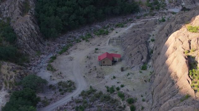 Drone footage reveals isolated wooden cabin with red metal roof nestled in rocky canyon of Sierra Madre Occidental mountains in Durango, Mexico. Dirt road winds through rugged terrain at golden hour.