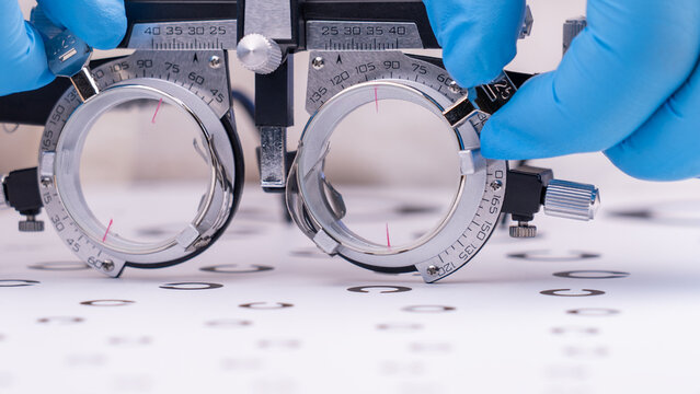 Close-up of a stereoscope. Blue-gloved hands adjust the device. Measurement markings cover the lenses. Letter chart sits below. This shows an eye examination process