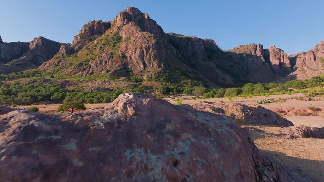 Aerial drone footage captures majestic rugged mountains of Sierra Madre Occidental with red rocky peaks, sparse green vegetation, arid sandy terrain, and clear blue skies during golden hour light.