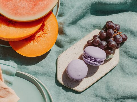 Overhead flat lay of summer picnic spread featuring slices of watermelon, cantaloupe, grapes, and lavender macarons on a sage green tablecloth