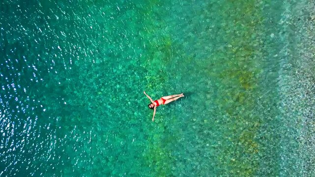 Woman in a red swimsuit lays on her back in the clear sea, top down view