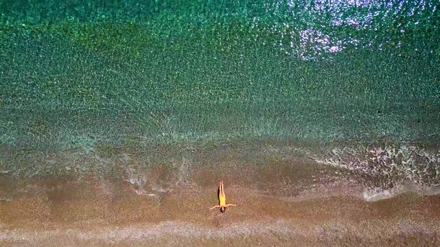 Woman in yellow swimsuit lays on a sandy beach, top down view. Vertical