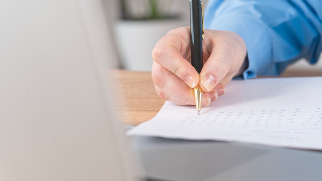 Close-up of a hand holding a black pen. Hand writes on a white document with neat script. Person wears a blue shirt, seated at a desk. Laptop screen is blurred in the background
