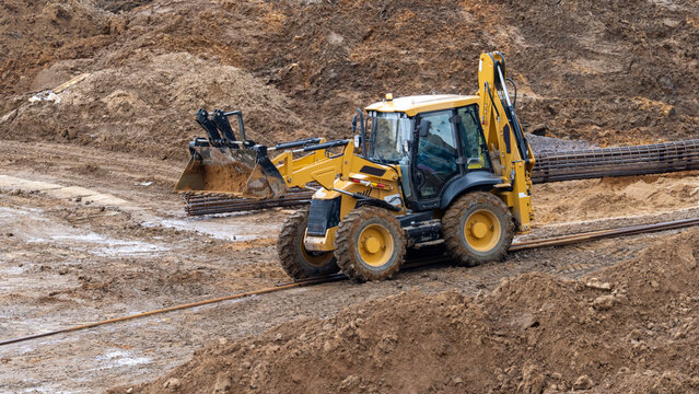 Yellow backhoe loader digs into muddy terrain. Its large tires sink into wet earth. The machine moves soil for land development. Construction site activity is clearly shown