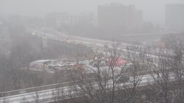 View of traffic moving along a highway during a heavy snowstorm with bare trees and buildings in the background in Tysons, Virginia, United States.