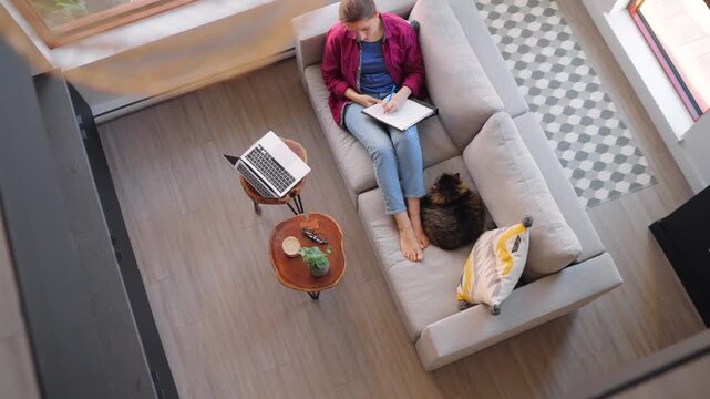 Overhead view of woman writing notes, lying on a couch at home