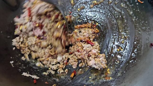 Stir frying minced pork with dried chili flakes and sauce in hot wok close up, spicy meat cooking process in Asian street food kitchen with oil bubbling and spatula mixing ingredients over high heat