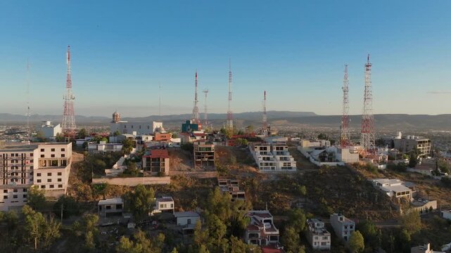 Aerial drone footage captures multiple telecommunication towers rising above residential hillside neighborhood in Durango, Mexico. Golden hour light illuminates buildings and mountains in background.