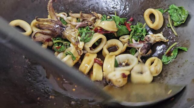 Stir frying squid rings and tentacles with basil leaves chili and sauce in hot wok close up, seafood cooking process in Asian street food kitchen with oil sizzling and ingredients mixing
