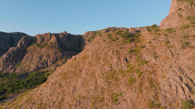 Stunning aerial footage of the Sierra Madre Occidental mountain range in Durango, Mexico. Rugged rocky peaks with sparse green vegetation bathed in warm golden hour sunlight against a clear blue sky.