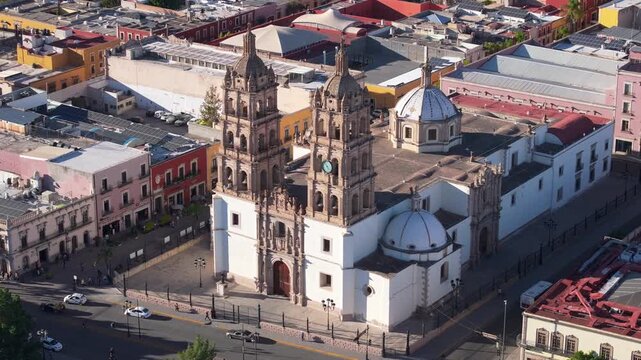Aerial drone footage captures the stunning baroque cathedral with ornate twin bell towers and blue domes in Durango Mexico. Colorful colonial buildings surround the historic church in the city center.