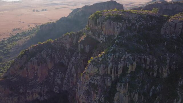 Aerial drone footage captures the rugged rocky cliffs and dramatic escarpments of a mountainous landscape bathed in warm golden hour sunlight, with sparse green vegetation clinging to steep slopes.