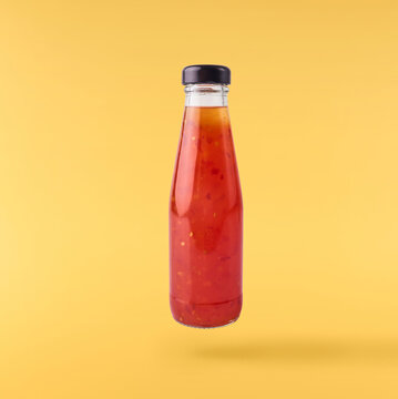 Levitating glass bottle of sweet chili sauce isolated on a solid yellow background. Minimalist studio food photography concept with zero gravity effect.