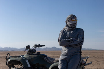 Man wearing a keffiyeh and sunglasses stands next to an ATV in a desert landscape. Adventure travel and outdoor activity concept. © Анна Маркина