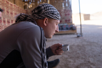 Fototapeta premium Man wearing a keffiyeh and grey jacket sits holding a small cup. Traveler enjoying a hot drink in a traditional bedouin tent.