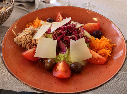 Mixed salad with tuna and purple radicchio served in a clay bowl. It also includes tomatoes, black olives, grated carrots, and is sprinkled with Parmesan or Pecorino cheese flakes.