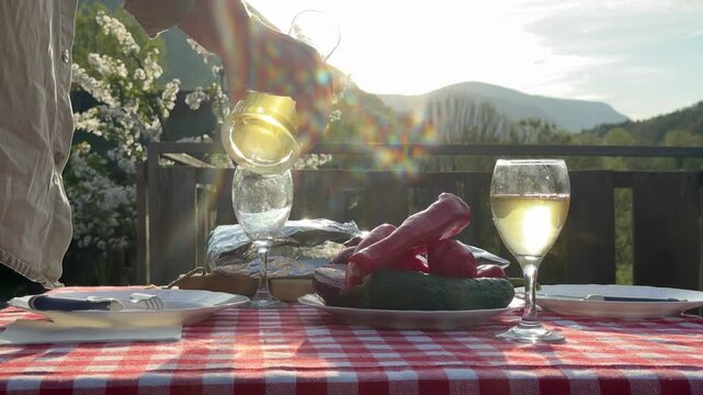 Close-up of a person pouring a drink into glasses at a rustic picnic table outdoors. Fresh vegetables, natural light, and countryside setting create a peaceful slow living atmosphere. Ideal for concep