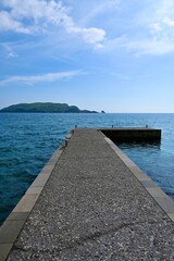 Obraz premium Perspective view of a stone pier extending into the calm blue sea under a bright sky, peaceful coastal landscape.