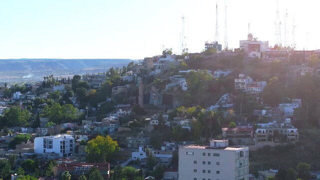 Red cable car gondola travels up steep hillside past white buildings and telecommunication towers in Durango, Mexico. Colonial church bell tower and residential homes dot the sunlit mountainside.