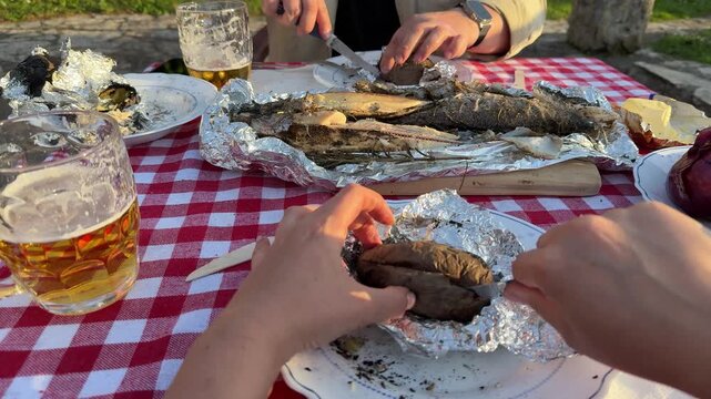 POV close-up shot of hands cutting baked potato wrapped in foil at an outdoor picnic table. Traditional rustic meal with grilled fish, beer, and homemade food. Warm sunset light and countryside atmos