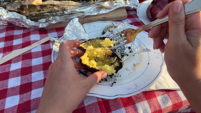 POV close-up shot of hands cutting baked potato wrapped in foil at an outdoor picnic table. Traditional rustic meal with grilled fish, beer, and homemade food. Warm sunset light and countryside atmos