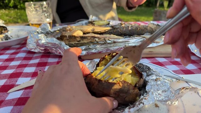 POV close-up shot of hands cutting baked potato wrapped in foil at an outdoor picnic table. Traditional rustic meal with grilled fish, beer, and homemade food. Warm sunset light and countryside atmos