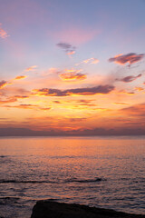 Fototapeta premium Vibrant sunset over calm ocean waters with colorful clouds reflecting on the surface, rocky shoreline visible in the foreground and distant mountains on the horizon