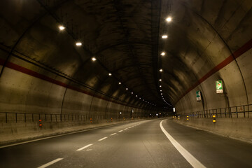 Fototapeta premium Dark concrete tunnel with illuminated lights and curved road, featuring road signs and safety barriers along the sides, creating a dramatic perspective view