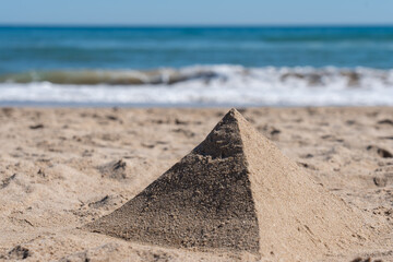 A perfectly shaped sand pyramid stands on a sunny beach, with soft ocean waves and a blue sky in the blurred background. © Sergej