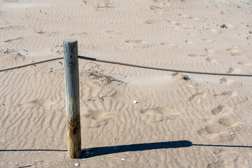 A weathered wooden post with a rope barrier stands in the sand, casting a long shadow on a sun-drenched beach. © Sergej