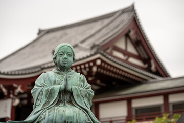 Fototapeta premium Tokyo, Japan - April 04, 2026: Zojo-ji Temple in Tokyo with a bronze Buddha statue in the foreground and the iconic red wooden beams and roof structure, Tokyo Tower rising in the background.