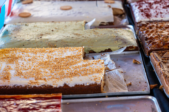 A variety of freshly baked tray cakes, including carrot cake with nuts and pistachio cream, displayed on metal trays.