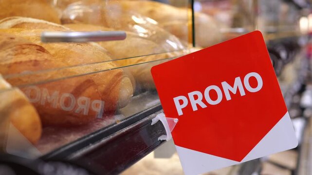 Red promotion sign hanging on a glass display case inside a bakery or supermarket, with fresh loaves of crusty bread visible on the shelf behind, signaling a discount or special offer