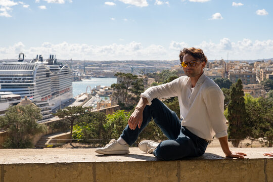 Young man in sunglasses sitting on a historic wall with a giant cruise ship in the background. Mediterranean travel concept in Valletta, Malta.