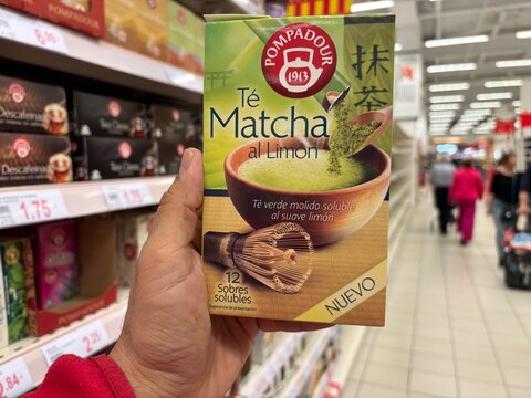 Alicante, Spain - April 25, 2026: Hand holding Pompadour Matcha Tea with Lemon packaging in a grocery store aisle, various tea products visible in the background