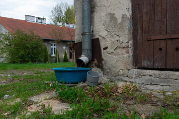 Fototapeta premium Old metal rain gutter on weathered wall draining into a blue plastic tub beside rustic buildings and grass. Useful for rainwater collection, rural yard, drainage system, old architecture and household