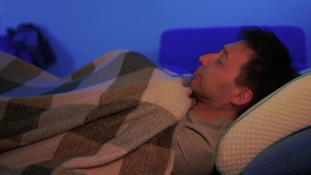 Worker sleeps peacefully. Individual lying awake under blanket. Adult reclines quietly on sofa in dim blue lighting. Peaceful scene of person resting in softly lit living room