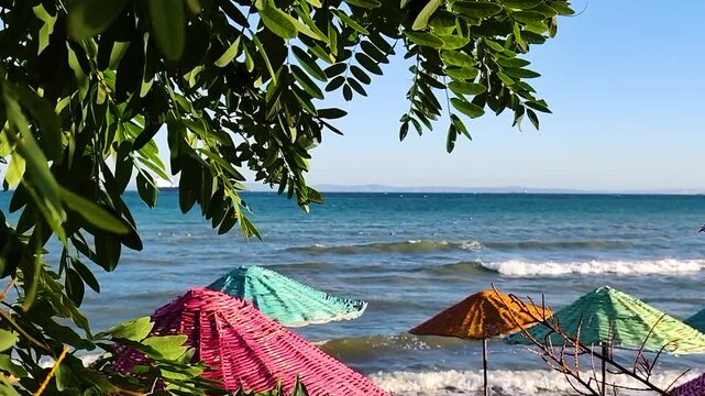 Vibrant Pink and Blue Wicker Sun Umbrellas on a Sunny Beach with Calm Sea Waves and Green Tree Leaves in Foreground - Summer Vacation and Travel Concept