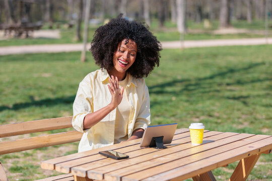 African woman waves and smiles while using a tablet in a park during a sunny day