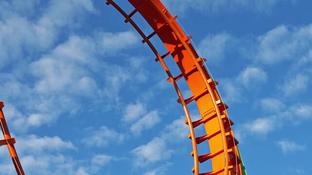 First person view moving along a thrilling orange roller coaster track with twists, turns, and loops against a sunny blue sky