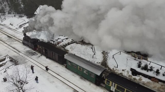 Historic steam locomotive with passenger cars pulling into a snow-covered rural station in Maramures, Romania, with thick smoke and winter scenery.