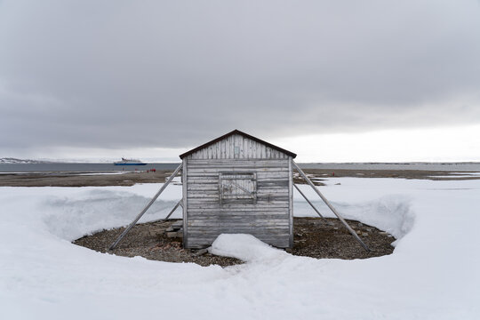 Svalbard, Svalbard and Jan Mayen - 21 June 2025: View of Kinnvika Station, a weathered wooden hut surrounded by snow and gravel under an overcast sky with a ship in the distance.