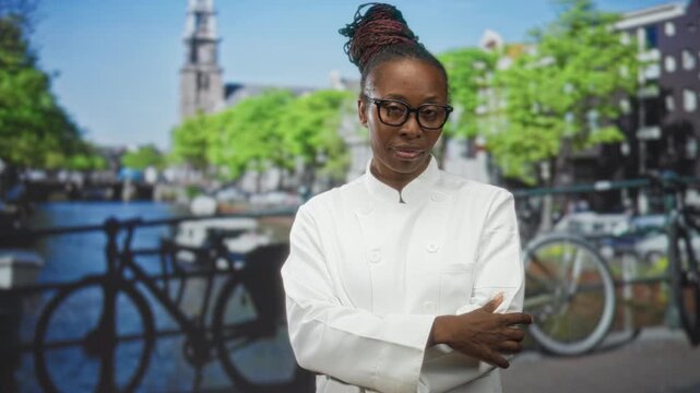 Woman chef in white uniform folds arms with hand on elbow in studio set with canal and bicycle backdrop; thoughtful resilience.
