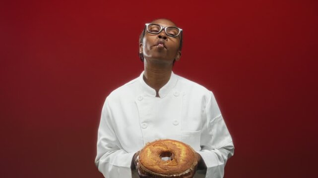 Woman cook in white chef coat and glasses holds a ring cake with bare hands and puckers lips while posing in a studio; playful confidence.