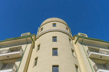 Obraz premium Low-angle view of a yellow building featuring a central rounded tower, decorative balconies, and green roof details, set against a vibrant, clear blue sky on a bright, sunny day