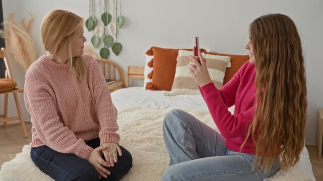 Two women sit crosslegged on a bed wearing sweaters as daughter shows a smartphone screen to mother in a bright bedroom, smiling and sharing; warmth connection.