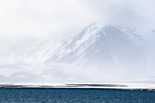 Svalbard, Svalbard and Jan Mayen - 24 April 2026: View of snow-capped mountains partially obscured by thick fog and low clouds above the icy waters of the Arctic coastline.