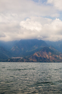Calming lake reflects distant, mist-covered mountains. Clouds drift above rugged, green-hued peaks. Water ripples gently under soft, overcast skies. Lake Atitlan, Guatemala.